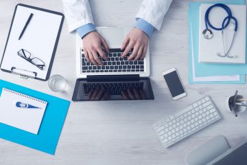 A clipboard with a pen and glasses on it, a notebook with a thermometer, a glass of water, a phone, a spare keyboard, a few files with a stethoscope on top of them, a pen holder and a medical professional's hands shown working on a laptop