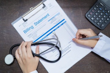 A billing statement being written on using the right hand while the left hand holds a stethoscope. A calculator is also shown on the right.